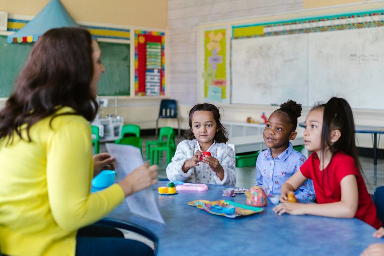 Three diverse young girls engage with a teacher in a vibrant classroom environment.
